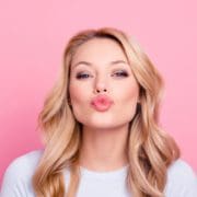 A woman with long blonde hair and light makeup is puckering her lips as if to blow a kiss. She is wearing a light top and is posed in front of a pink background, radiating confidence with help from Beauty Boosters by Aesthetics By Boss.