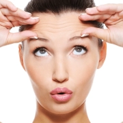 A woman pressing her fingers against her forehead, raising her eyebrows and pursing her lips to reveal visible forehead wrinkles—an expressive look that highlights the need for Beauty Boosters or Anti Wrinkle Injections; the background is white.