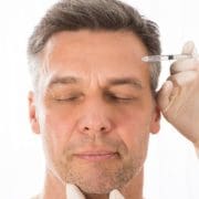 A man with closed eyes receives an injection near his temple from a gloved hand, suggesting a cosmetic procedure such as Anti Wrinkle Injections or Beauty Boosters.