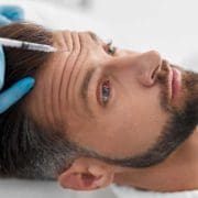 A man lies calmly as he receives anti wrinkle injections to his forehead from a professional wearing blue gloves. He gazes upward while the Aesthetics By Boss procedure is being carefully performed.