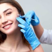 A young woman with long brown hair smiles as a person wearing blue medical gloves prepares to give her Anti Wrinkle Injections with a syringe. The background is softly blurred.