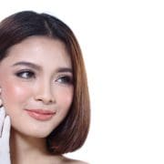 A woman with short brown hair smiles slightly as a gloved hand administers an Anti Wrinkle Injection to her cheek. The background is white, and the woman appears calm and relaxed, embracing subtle beauty boosters.