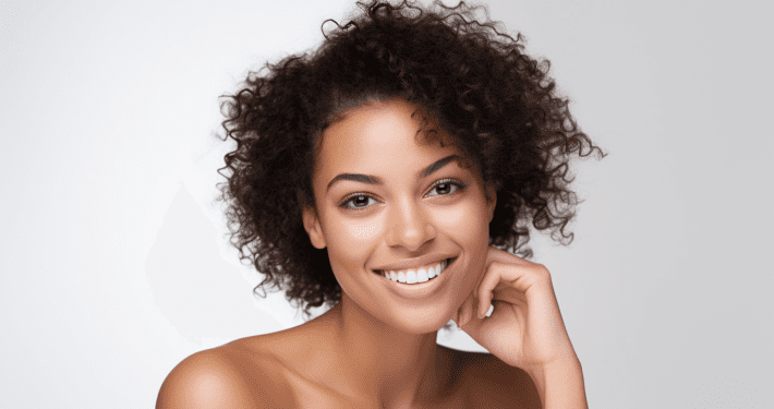A young woman with curly hair smiles warmly at the camera, resting her chin on her hand. She has clear skin thanks to Beauty Boosters and is posed against a soft, light gray background.